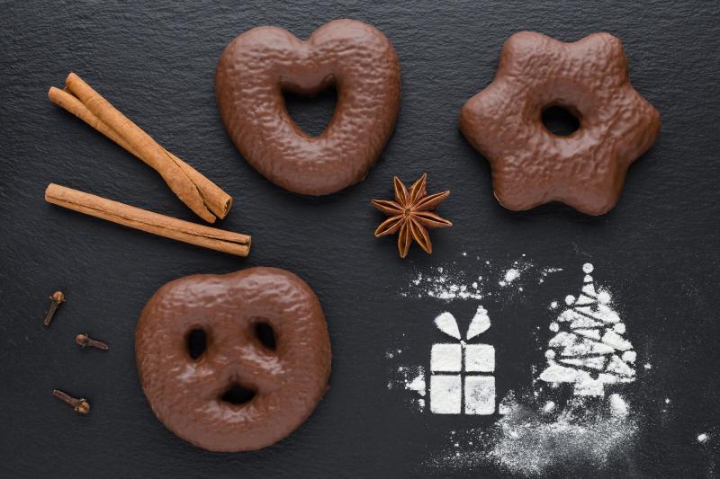 Dunkle Unterlage. Darauf wenige Zimtstangen und Gewürznelken; aus Staubzucker mit Schablone ein Packerl und ein Christbaum; 3 Schoko-Lebkuchen: Breze, Herz, Stern