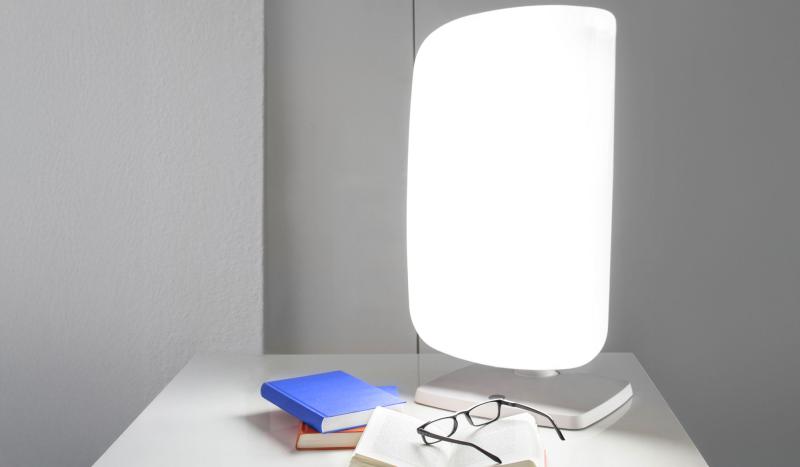 Auf dem Bild ist eine eingeschaltete, rechteckige Tageslichtlampe auf einem weißen Tisch zu sehen. Sie leuchtet hellweiß und steht vor einer hellgrauen Wand. Neben der Lampe liegen zwei Bücher, darauf eine Brille. Die Szene wirkt ruhig, minimalistisch und erinnert an einen Arbeitsplatz oder Nachttisch.