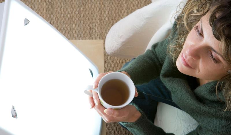 von oben: lockige Frau mit Teehäferl in Händen sitzt mit geschlossenen Augen vor einer eingeschalteten Tageslichtlampe