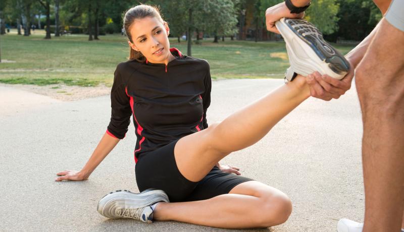 Junge, schwarz gekleidete, dunkelhaarige Frau mit Pferdeschwanz sitzt draußen auf dem Boden und lässt sich mit ausgestrecktem Bein von einer zweiten - kaum sichtbaren - Person die Wade strecken.