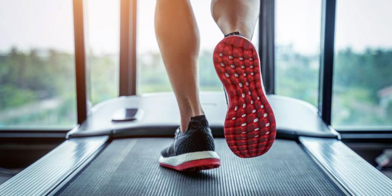 Nahaufnahme von laufenden Beinen auf einem Laufband. Der Fokus liegt auf der roten Sohle eines Sportschuhs während des Schritts nach vorne. Durch die großen Fenster im Hintergrund fällt Tageslicht, draußen ist eine grüne Landschaft zu sehen. Die Szene vermittelt Bewegung, Training und Ausdauer.