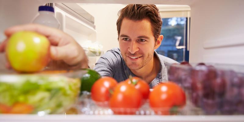 Der Blick in einen geöffneten Kühlschrank zeigt frische Lebensmittel: vorn liegen rote Tomaten und Weintrauben, daneben eine Salatschüssel und Limetten. Eine Person greift von außen nach einem grünen Apfel. Im Hintergrund stehen weitere Vorräte wie eine Milchflasche. Die Szene vermittelt den Moment der Auswahl eines Snacks, während die Farben der Zutaten Vielfalt und Frische betonen.