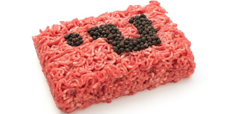 Ein rechteckiger Block aus rohem Faschiertem zu sehen, der auf einer hellen, neutralen Fläche liegt. Das Fleisch ist frisch und rötlich gefärbt, mit deutlich erkennbarer, grob gewolf­ter Struktur. Auf der Oberseite sind schwarze Pfefferkörner so angeordnet, dass sie ein Fragezeichen ergeben.