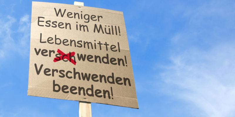 Auf dem Foto ist ein Protest- oder Infoplakat aus Karton zu sehen, das vor einem hellblauen Himmel hochgehalten wird. Auf dem Schild steht in schwarzer Schrift: „Weniger Essen im Müll! Lebensmittel ver(s)chwenden! Verschwenden beenden!“ Das Wort „verschwendet“ wurde handschriftlich mit roter Farbe korrigiert. Das Bild vermittelt eine klare Botschaft gegen Lebensmittelverschwendung.