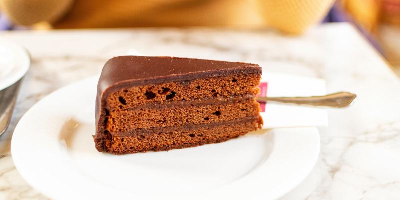 Das Bild zeigt ein einzelnes Stück Schokoladentorte auf einem weißen Teller. Die Torte besteht aus mehreren dunklen, gleichmäßig geschichteten Lagen und ist vollständig mit glatter Schokoglasur überzogen. Im Hintergrund liegt ein Löffel, leicht unscharf, und die Szene befindet sich auf einer hellen Marmoroberfläche in warmem Umgebungslicht.