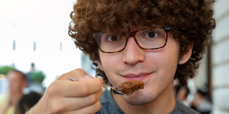 Junger Mann mit lockigem, voluminösem Haar und mit Sachertortenstück auf Kuchengabel, die zum Mund geführt wird. Die Szene wirkt wie in einem Café oder Außenbereich, da im Hintergrund unscharfe Menschen und helle Gebäude zu sehen sind. Die Beleuchtung ist natürlich und erzeugt einen warmen Gesamteindruck.