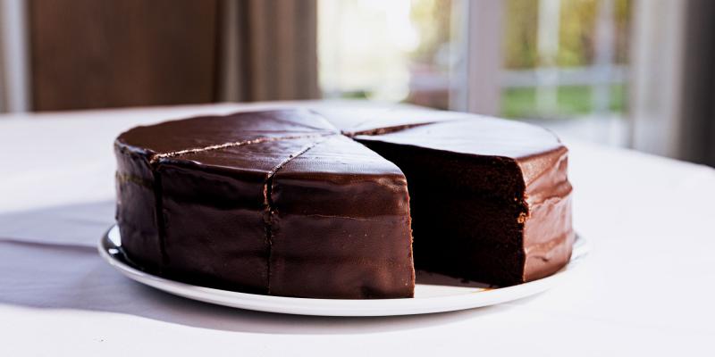 Auf einem weißen Teller steht eine runde Schokoladentorte, von der ein großes Stück herausgeschnitten wurde. Die Torte ist vollständig mit dunkler, glänzender Glasur überzogen und zeigt mehrere saftige Schichten im Anschnitt. Der Teller steht auf einer hellen Tischdecke, im Hintergrund sind Fenster und weiches Tageslicht zu sehen.