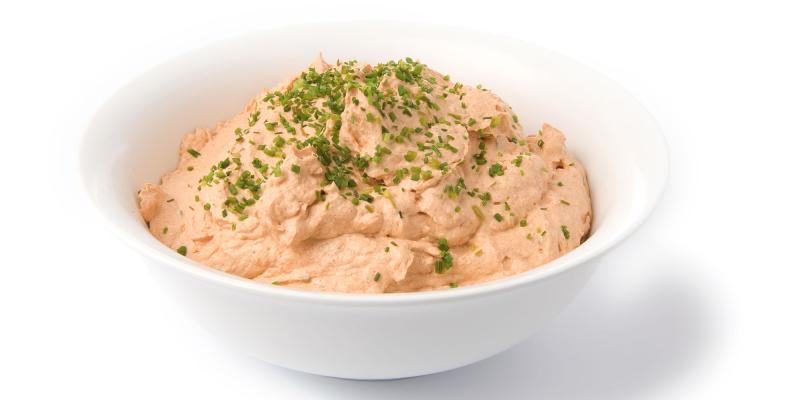 Das Bild zeigt eine weiße Schüssel, die mit einer cremigen, hellorangefarbenen Masse gefüllt ist. Die Konsistenz wirkt glatt, leicht aufgeschlagen und fluffig. Auf der Oberfläche der Creme sind fein gehackte grüne Kräuter – vermutlich Schnittlauch oder Petersilie – verteilt. Die Schüssel steht vor einem reinweißen Hintergrund, wodurch der Inhalt klar hervorgehoben wird.