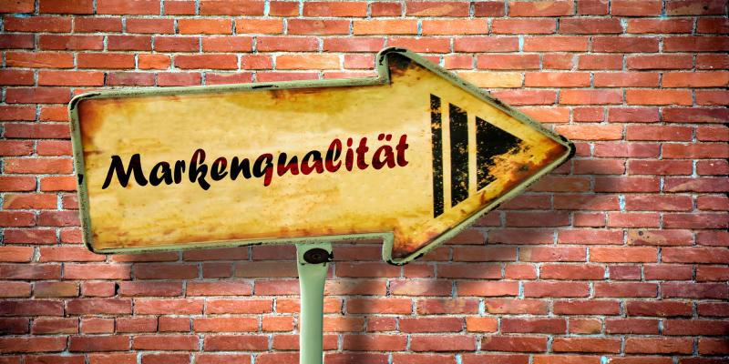 Ein gelbes, verwittertes Schild in Pfeilform zeigt nach rechts und steht vor einer roten Backsteinwand. Auf dem Schild steht das Wort „Markenqualität“ in schwarzer Schrift, daneben drei grafische Pfeile. Das Schild wirkt alt und leicht beschädigt, wodurch ein rustikaler, vintageartiger Eindruck entsteht.