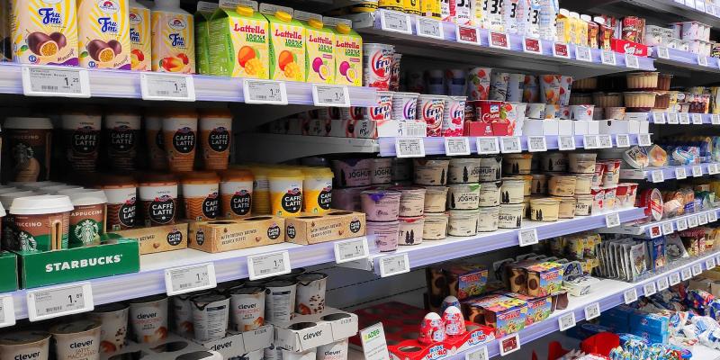 Ein Supermarktregal ist dicht gefüllt mit verschiedenen Lebensmitteln und Getränken. Zu sehen sind Kühlprodukte wie Joghurts, Desserts, gekühlte Kaffees sowie Fruchtgetränke und Snacks. Die Verpackungen sind bunt und vielfältig, ordentlich in Reihen sortiert. Preisetiketten befinden sich gut sichtbar unter jedem Produkt.