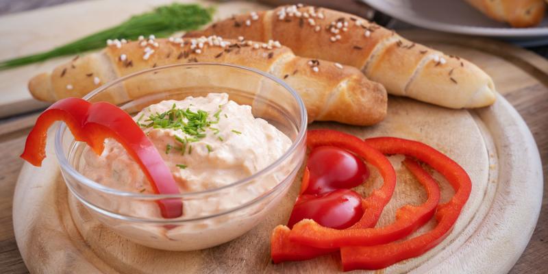 Auf einem runden Holzbrett steht eine kleine Glasschüssel mit einem cremigen, hellen Brotaufstrich. Der Aufstrich ist mit etwas Petersilie garniert. Am Rand der Schüssel steckt ein Stück rote Paprika. Neben der Schüssel liegen frische Paprikaringe und zwei kleine rote Tomaten. Im Hintergrund befinden sich mehrere längliche Gebäckstücke, sogenannte Salzstangerl.