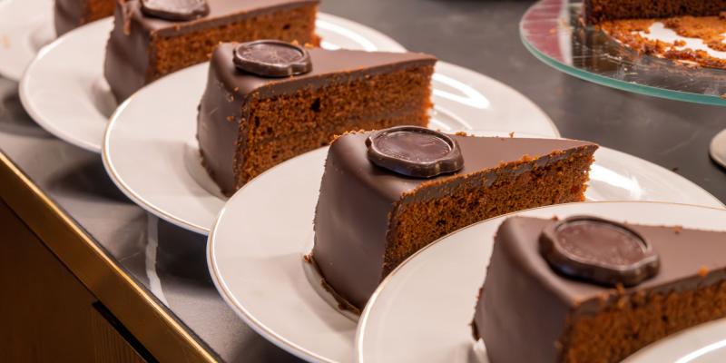 Das Bild zeigt mehrere Stücke einer schokoladenglasierten Torte (Sachertorte), die jeweils auf weißen Tellern in einer Reihe angeordnet sind. Jedes Tortenstück trägt oben eine runde Schokodekoration. Die Stücke stehen dicht beieinander auf einer Theke, im Hintergrund ist ein Glasservierteller mit weiteren Kuchenresten erkennbar.