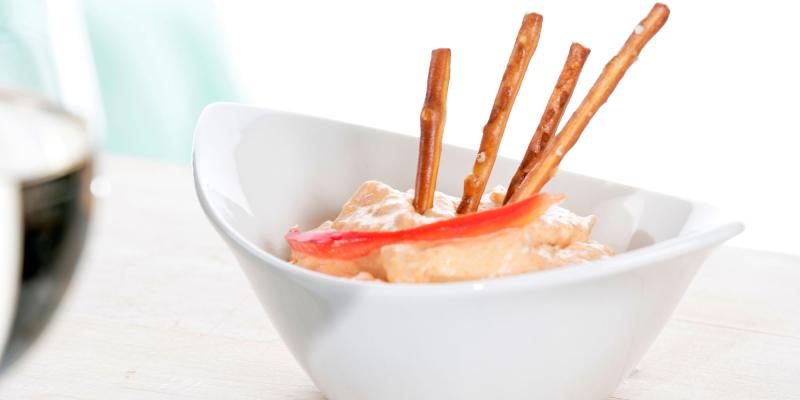In einer weißen Schüssel befindet sich ein cremiger, heller Dip. Aus dem Dip ragen mehrere dünne Salzstangen nach oben. Auf der Oberfläche liegt ein Streifen roter Paprika. Die Schüssel steht auf einem hellen Tisch. Im Hintergrund ist ein unscharfes Glas sehen.