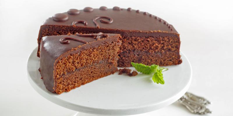 Das Bild zeigt eine runde Schokoladentorte auf einem weißen Kuchenständer. Ein angeschnittenes Stück ist leicht herausgezogen und gibt den Blick auf den dunklen, saftigen Teig mit mittlerer Schicht frei. Die Torte ist vollständig mit glänzender Schokoglasur überzogen und mit kleinen dekorativen Schokopunkten verziert. Daneben liegt ein frisches Minzblatt als Garnitur.