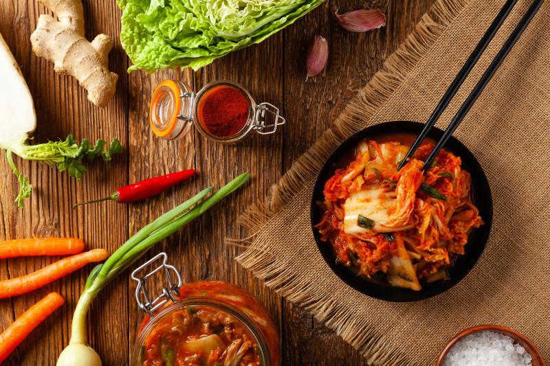 Kimchi auf Teller, dieser steht auf einem Jute-Tischtuch, daneben verschiedenes Gemüse, unter anderem Chinakohl, Karotten