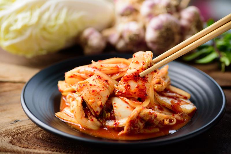 Kimchi auf schwarzem Teller, Stäbchen ergreifen eine Portion Kimchi, Chinakohl im Hintergrund