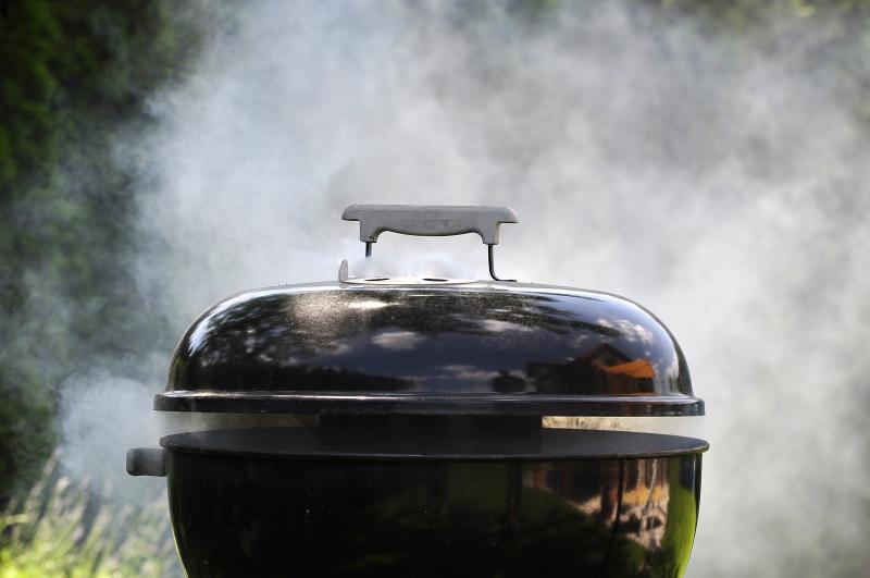 Schwarzer Kugelgrill mit geschlossenem Deckel, aus dem Lüftungsschlitz quillt dichter Rauch, dahinter grüne Sträucher im Garten.