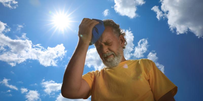 Das Bild zeigt eine Person im Freien unter einem blauen Himmel mit vereinzelten weißen Wolken. Die Sonne steht hell und strahlend im oberen linken Bereich des Bildes. Im Vordergrund hält die Person ein blaues Tuch an die Stirn, als würde die Person versuchen, sich abzukühlen. Die Person trägt ein kurzärmliges gelbes Shirt. Die starke Sonneneinstrahlung und die Geste mit dem Tuch deuten auf hohe Temperaturen hin.