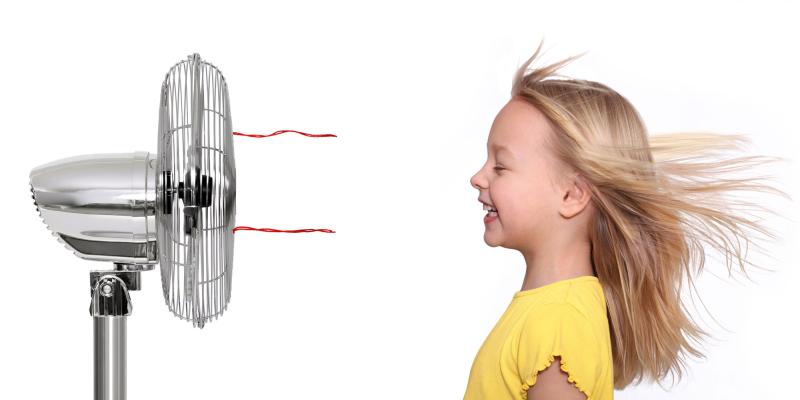 Das Bild zeigt eine Szene vor einem hellen, neutralen Hintergrund. Auf der linken Seite steht ein großer Ventilator mit sichtbarem Schutzgitter und rot angedeuteten Luftströmungen. Auf der rechten Seite befindet sich ein Kind mit langen, nach hinten wehenden Haaren, die vom Luftstrom bewegt werden. Das Kind trägt ein gelbes Oberteil und steht seitlich dem Ventilator zugewandt. Die Darstellung macht deutlich, wie der Ventilator für Abkühlung sorgt und Bewegung in die Haare bringt.