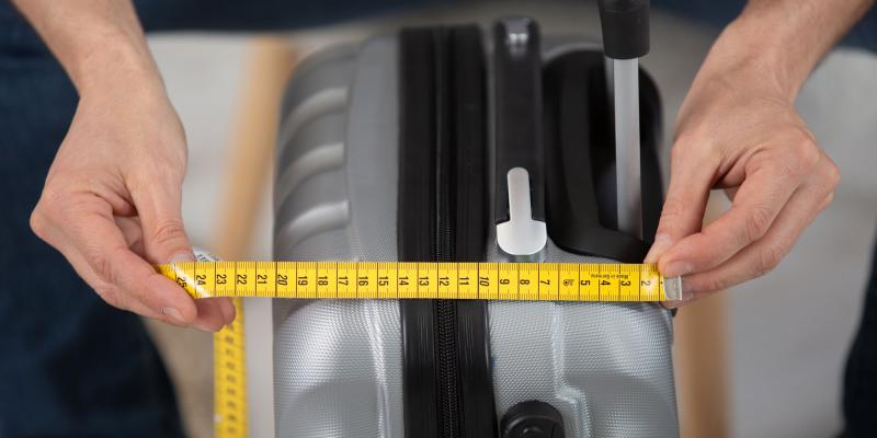 2 Hände halten ein gelbes Maßband über die Breitseite eines kleinen Hartschalenkoffers