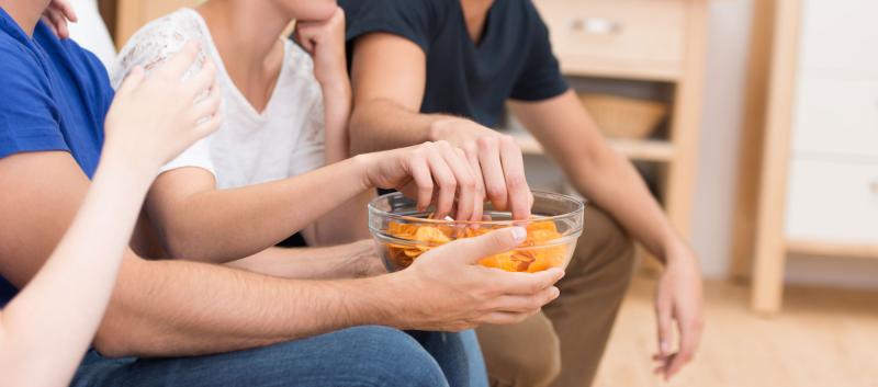 viele Hände von auf einem Sofa sitzenden Menschen greifen in eine Glasschüssel voll mit Kartoffelchips