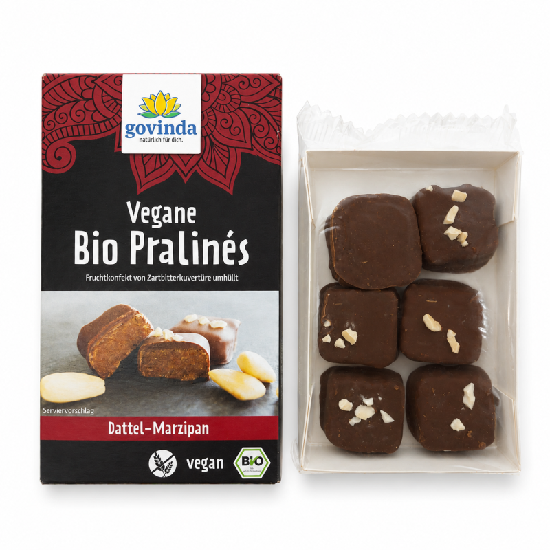 Geöffnete Packung „Govinda" Vegane Bio Pralinés „Dattel-Marzipan" neben der herausgenommenen Kunststoffschale mit sechs quadratischen, schokoladeüberzogenen Pralinen, einige mit Mandelstücken bestreut.