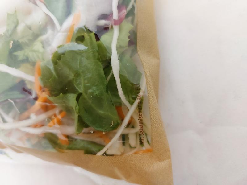Nahaufnahme einer verpackten Salatmischung mit grünen Blättern, Karottenstreifen und Weißkraut. Auf der Verpackung steht „Mit Salatmischung aus EU.“