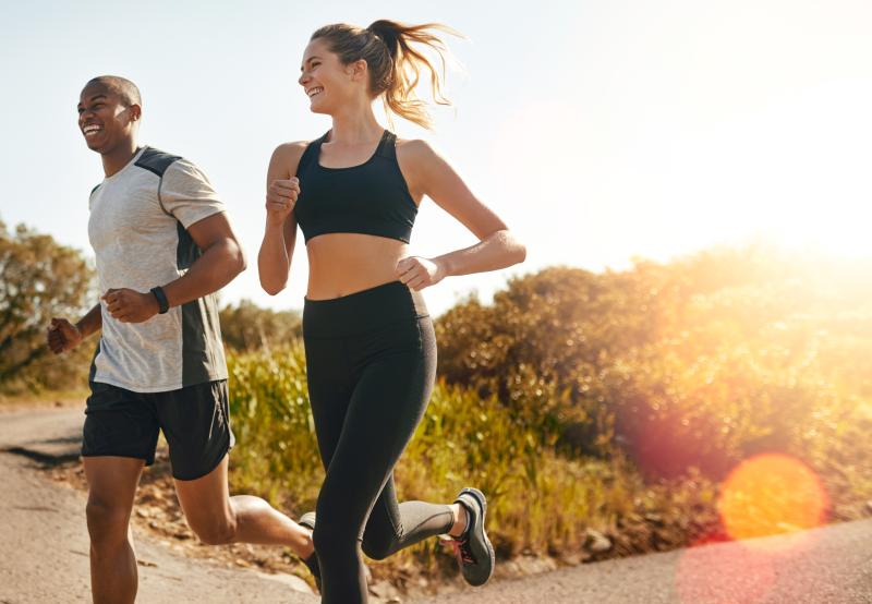 Zwei lächelnde Personen joggen bei sonnigem Wetter gemeinsam auf einem Weg in der Natur.
