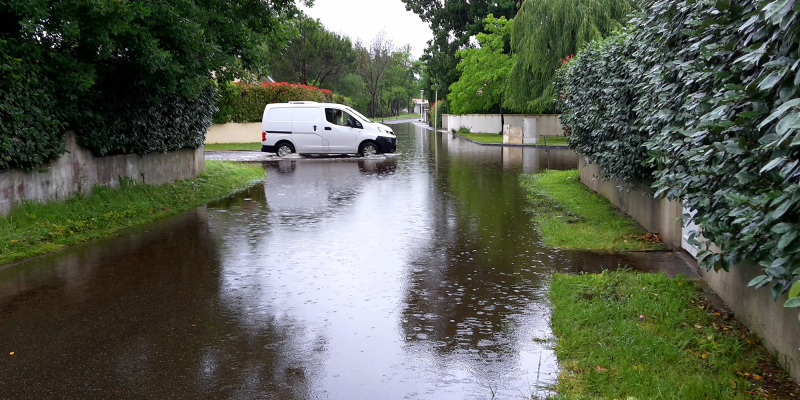 Eine überflutete Wohnstraße steht nach starken Niederschlägen unter Wasser. Die Fahrbahn ist großflächig überschwemmt und spiegelt Bäume, Hecken und den grauen Himmel wider. Im Hintergrund befindet sich ein weißer Lieferwagen, der nahe der überfluteten Stelle steht. Beidseits der Straße verlaufen Grünstreifen und dichte Hecken entlang von Grundstücksmauern. Die Szene zeigt die Auswirkungen eines Hochwasserereignisses in einem Wohngebiet.