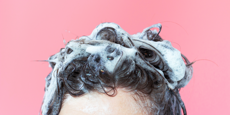 Nahaufnahme eines Kopfes während der Haarwäsche vor einem rosa Hintergrund. Das dunkle Haar ist mit reichlich weißem Schaum bedeckt, der sich in weichen Wellen und Locken über den oberen Kopfbereich legt. Zahlreiche kleine Bläschen und feuchte Strähnen sind sichtbar. Der Bildausschnitt zeigt nur den oberen Teil der Stirn und die Haare. Die Farbgebung aus Rosa, Weiß und Dunkelbraun erzeugt einen klaren, kontrastreichen und modernen Bildeindruck.