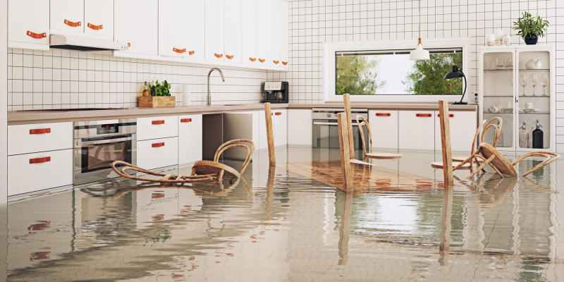Eine moderne Wohnküche ist infolge eines Hochwasserereignisses überflutet. Das Wasser bedeckt den gesamten Boden und reicht bis an Möbel und Kücheneinrichtung. Mehrere Stühle sind umgekippt oder teilweise im Wasser versunken. Weiße Küchenschränke, Arbeitsflächen und ein Esstisch bleiben sichtbar. Durch große Fenster fällt Tageslicht in den Raum und beleuchtet die spiegelnde Wasseroberfläche. Die Szene verdeutlicht erhebliche Wasserschäden in einem Wohngebäude durch Überschwemmung.