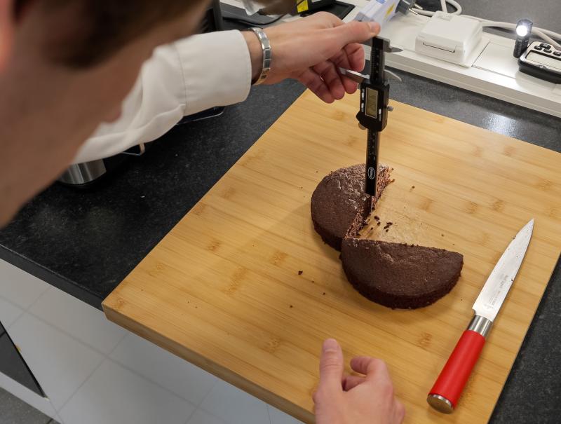 Eine Person im Laborkittel misst mit einem digitalen Messschieber die Höhe eines halbierten Schokokuchens auf einem Holzbrett. Daneben liegt ein Messer mit rotem Griff.