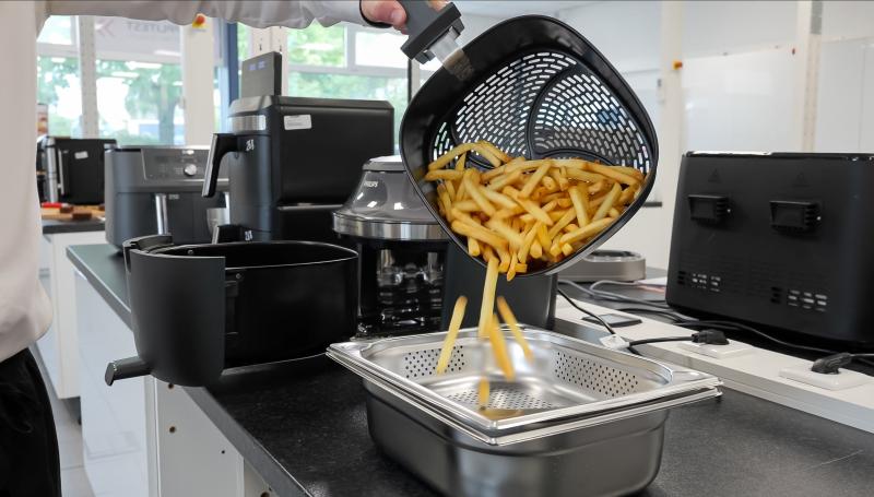 Eine Person schüttet fertige Pommes frites aus dem Korb einer „Philips" Heißluftfritteuse in eine perforierte Edelstahlschale. Im Hintergrund weitere Testgeräte.