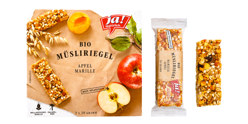 „ja! Natürlich" Bio-Müsliriegel „Apfel Marille", Faltschachtel zu 3 x 30 Gramm, daneben ein verpackter und ein ausgepackter Riegel. Die Verpackung nennt „rein pflanzlich", „Ballaststoffquelle" und „palmölfrei".
